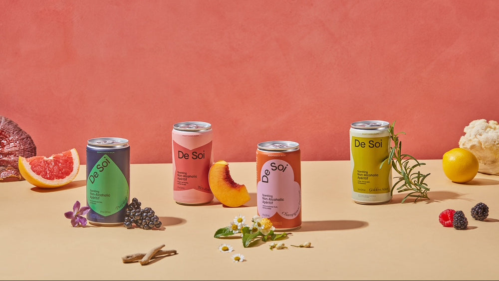 Four cans of 'De Soi' drinks on a pink background with fruits and herbs.